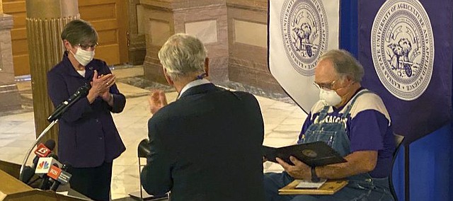 In this Tuesday, May 5, 2020 photo provided by the State of Kansas, Kansas Gov. Laura Kelly, left, and Kansas State University President Richard Myers award a bachelor's degree to Dennis Ruhnke, right, in Topeka, Kansas. The degree was bestowed upon Ruhnke, a retired Kansas farmer who shipped one of his five N-95 masks to New York Gov. Andrew Cuomo despite worries amid the coronavirus outbreak for his wife who has one lung. Kelly said that Ruhnke "perfectly encapsulates K-State's democratic mandate to be of service to people." (State of Kansas via AP)